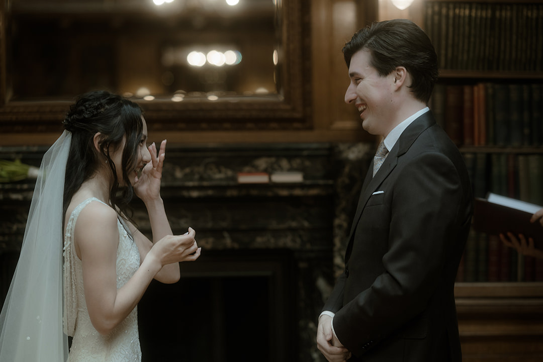 A side-profile view of a bride in a beaded white gown and long veil wiping away a happy tear as she faces her groom, who stands in a dark suit and patterned tie, smiling warmly. Behind them, a marble fireplace and rows of leather-bound books line the wood-panelled library walls of New Library at the Royal College of Physicians in Edinburgh.
