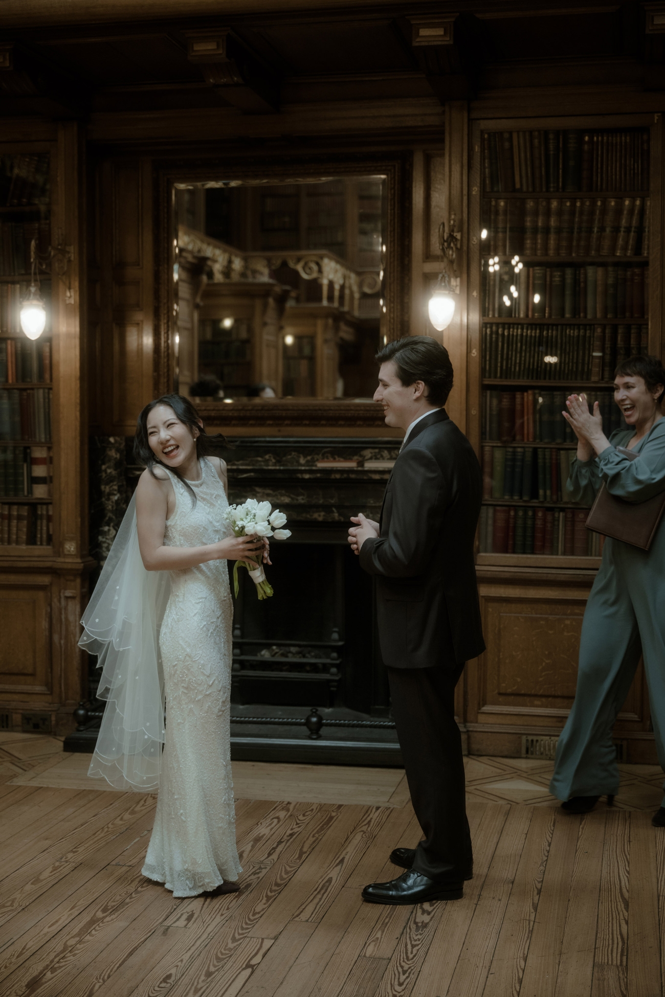 The bride laughs joyfully, clutching a small bouquet of white tulips, as the groom looks on and the humanist celebrant applauds behind them. The pair stand before a marble fireplace flanked by floor-to-ceiling bookshelves.
