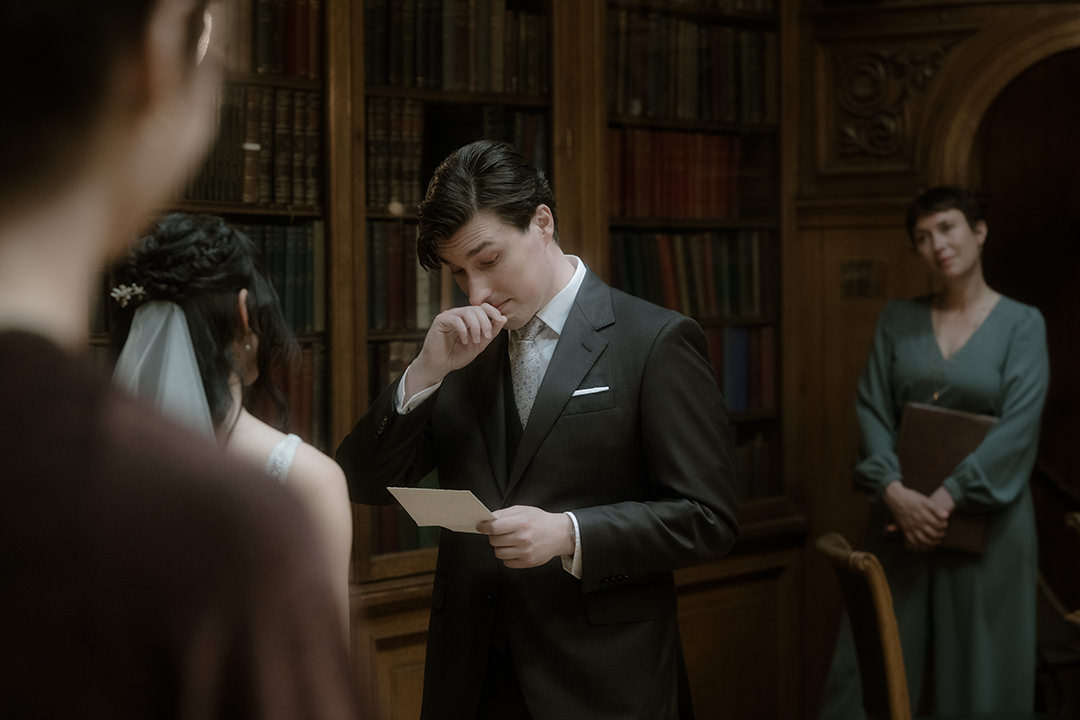 A close-up of the groom wiping his eye with the back of his hand while holding his vow card, his face turned slightly downward.