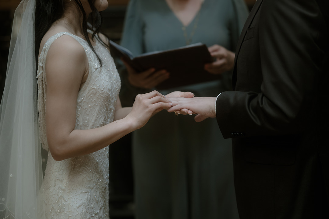 A tight shot of the bride’s hands as she gently slides a wedding band onto the groom’s finger. The textured beading on her dress and the celebrant’s open book in soft focus behind.
