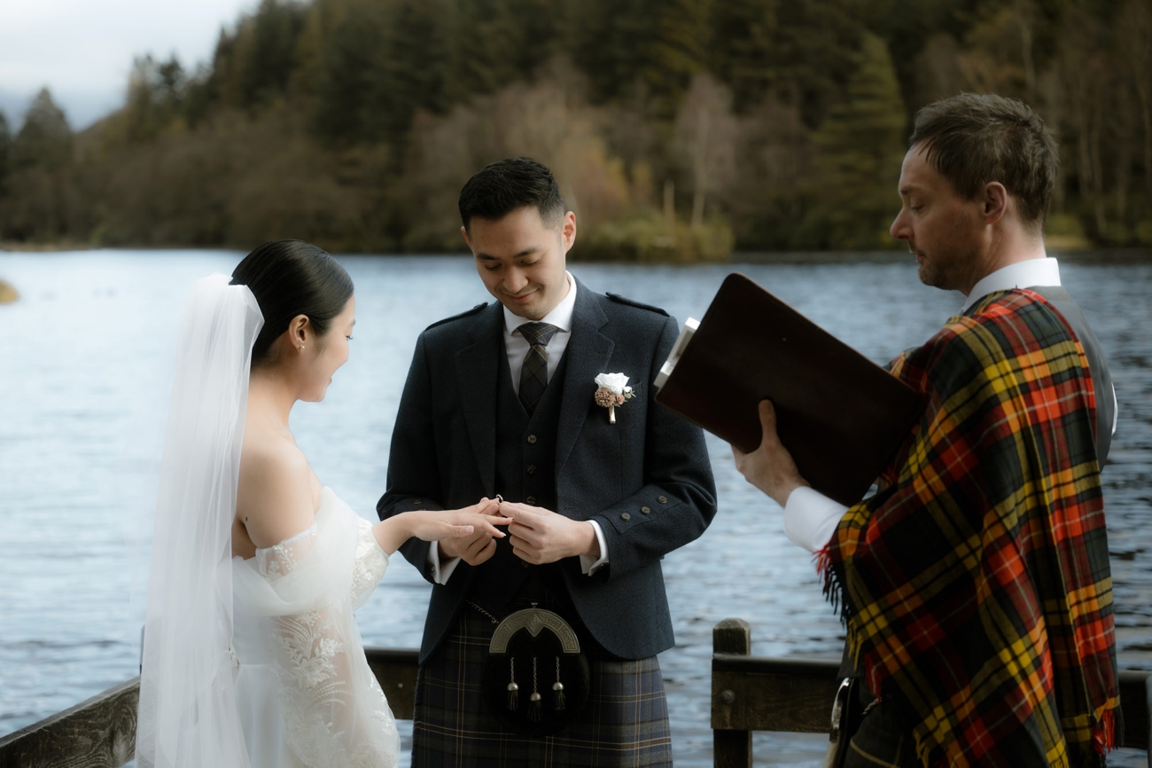 Ring exchange: the groom slides a ring onto the bride’s hand, a kitled humanist celebrant observes.