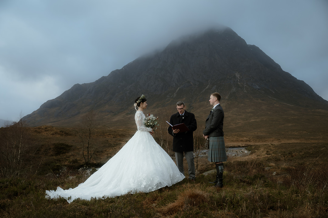A wide‐angle shot of a humanist wedding on a windswept Highland moor. The bride, in a full-skirted white lace ballgown and flower crown, stands facing her groom—dressed in a grey jacket, tartan kilt, and knee-high socks—while the celebrant reads from a leather-bound book.