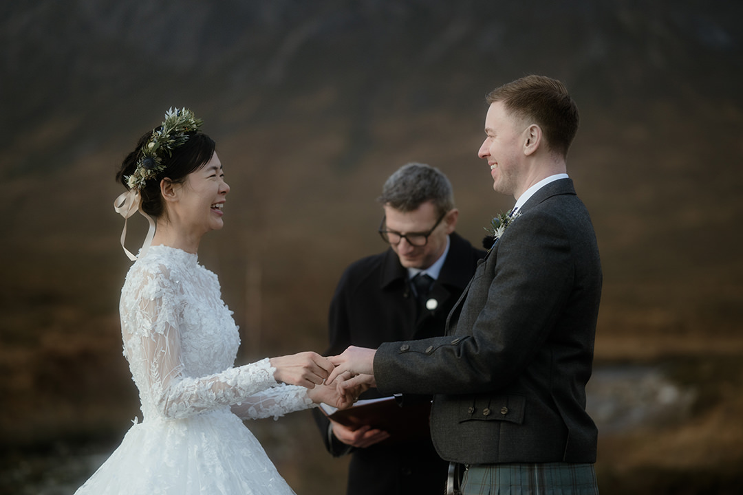 The bride’s delicate lace sleeves and floral wreath accent her bright laughter as she slips a ring onto the groom’s hand