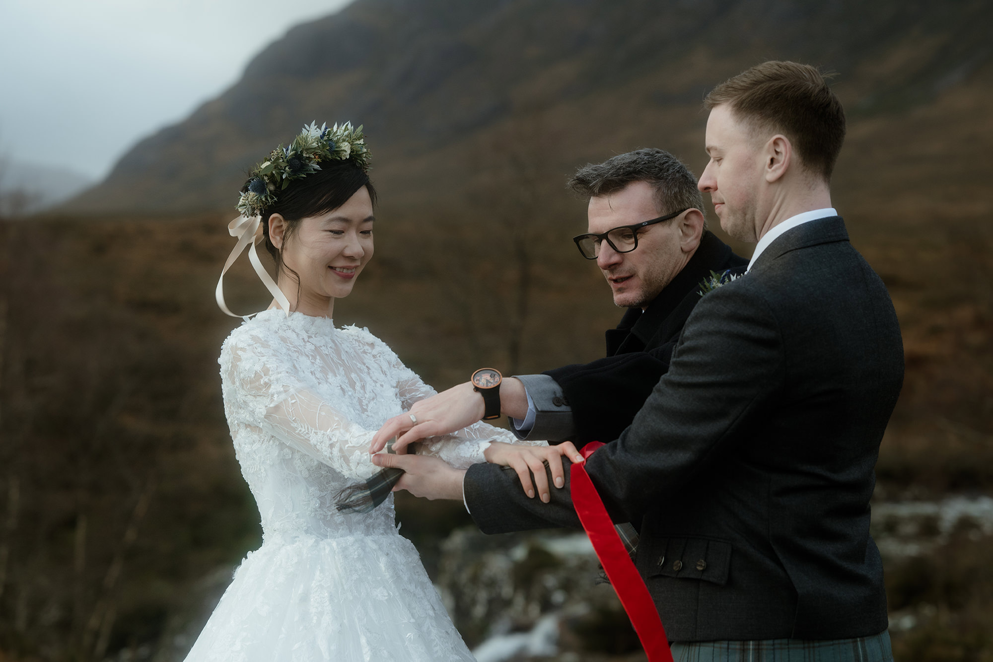 A closer view of the handfasting ritual. The celebrant wraps a length of red ribbon around the couple’s joined wrists, his hands guiding the bride’s and groom’s arms together.