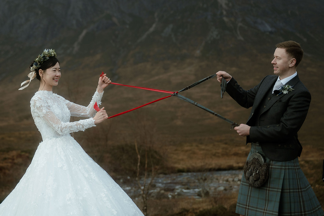 A dynamic mid-shot of the bride and groom each holding one end of two braided ribbons—she grips bright red cords, he holds darker ones—playing out the symbolic 'tying the knot'