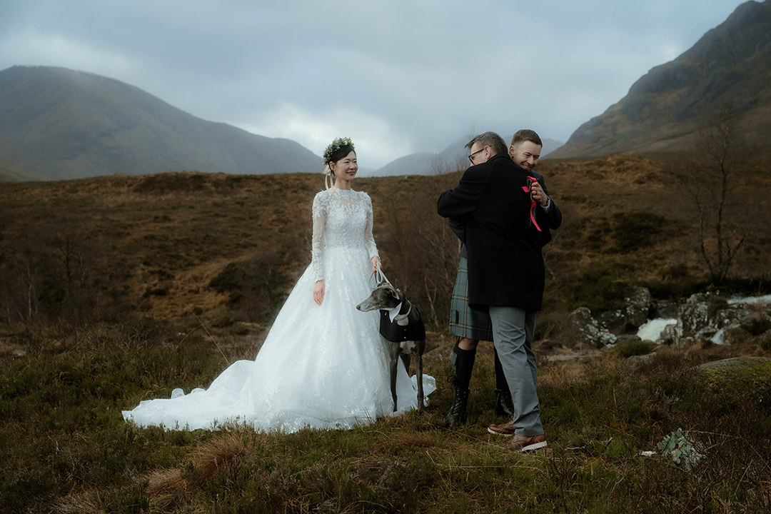 A celebratory moment after the ceremony: the groom embraces the officiant in a warm hug as the bride stands beside them, her train fanned on the grass. She holds the leash of a sleek greyhound wearing a little bowtie