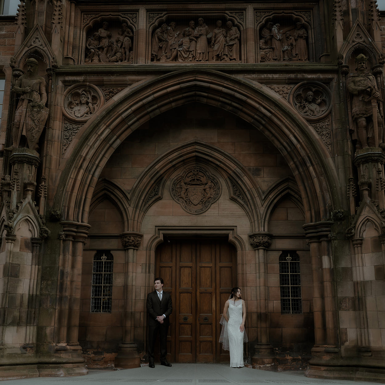 A bride and groom stand apart on either side of a grand Gothic archway carved in sandstone, each gazing toward the other against the backdrop of a large wooden doorway and sculpted figures above.