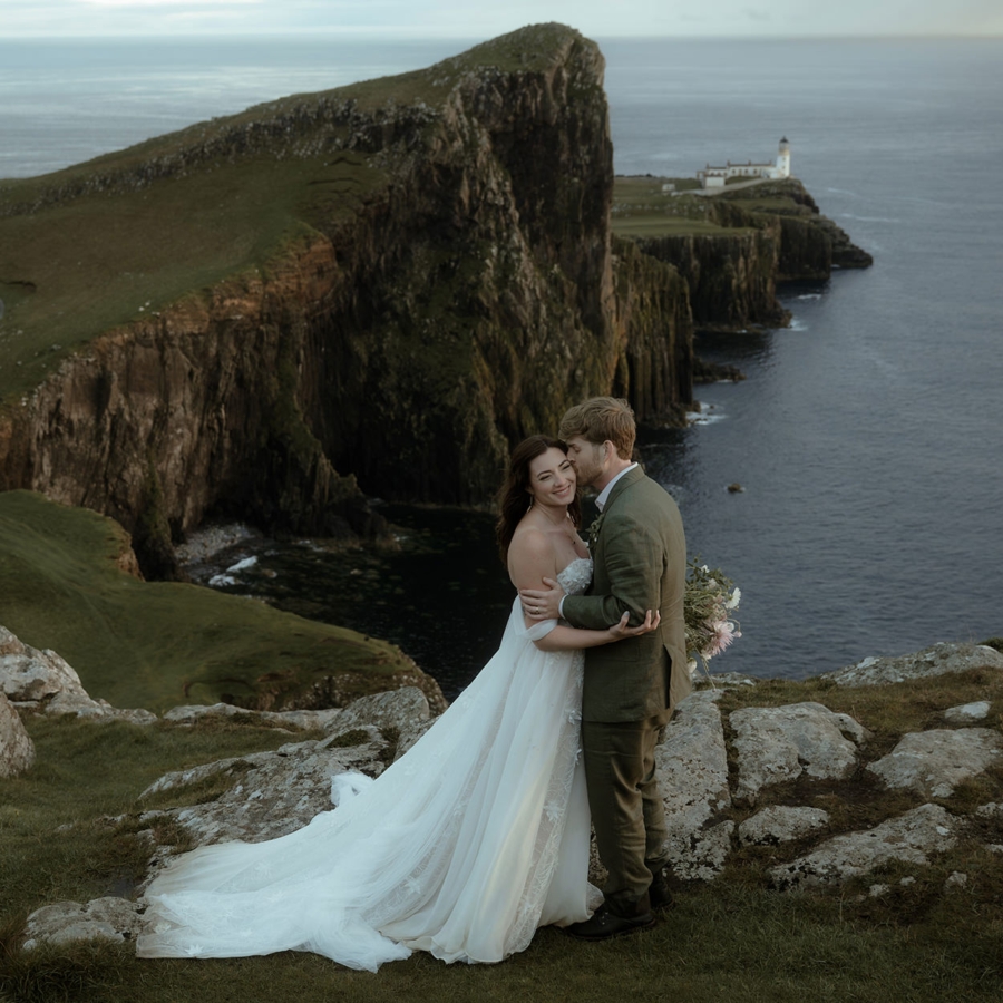 Bride and groom embrace on the cliffs at Neist Point, Isle of Skye, with the lighthouse and sea behind—flowing lace dress, green suit, soft evening elopement light