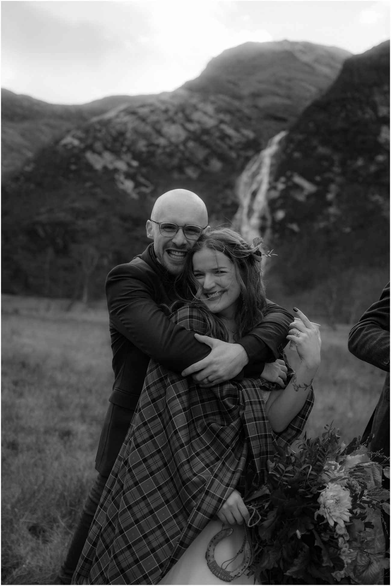 Steall Falls elopement in Glen Nevis 13 The groom wraps his arms tightly around the bride in an affectionate hug. Her eyes sparkle as she smiles at the camera, tartan cloak draped over her shoulders