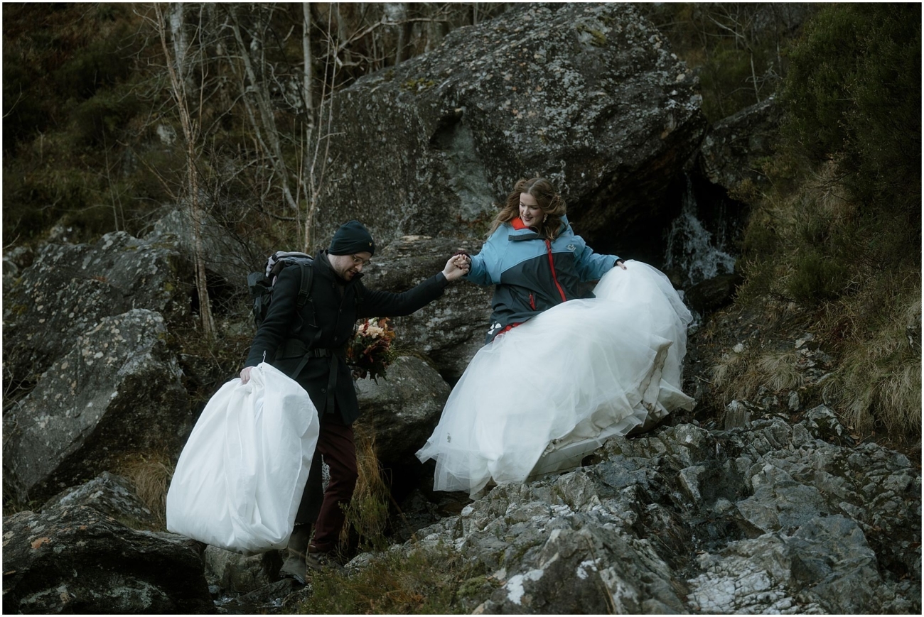 Steall Falls elopement in Glen Nevis 3 The groom helps the bride descend a rocky path near a small waterfall. She lifts her voluminous white dress as he steadies her, both dressed warmly for the hike