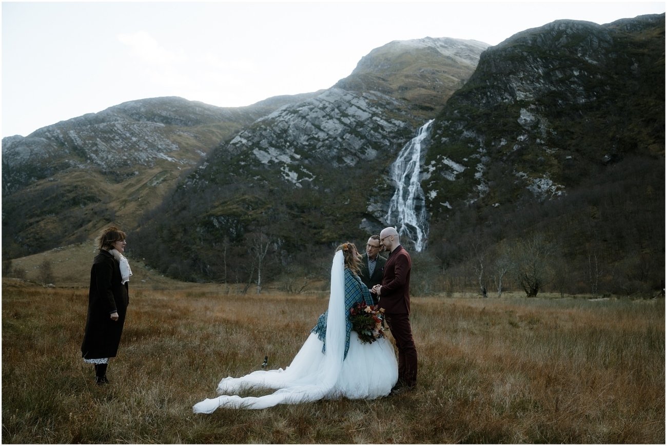 Steall Falls elopement in Glen Nevis 7 The ceremony begins. The bride and groom stand in the grassy field with Steall Falls behind them. Her veil trails out dramatically as the celebrant stands opposite