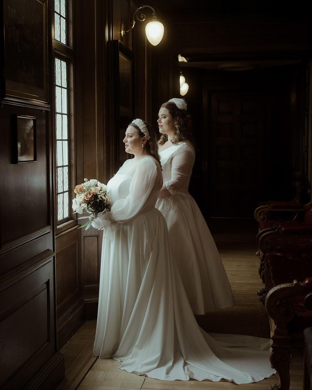 Home 19 Two brides in long white gowns stand side by side in a wood-paneled hallway, softly lit by wall sconces on their wedding day at Edinburgh New Library