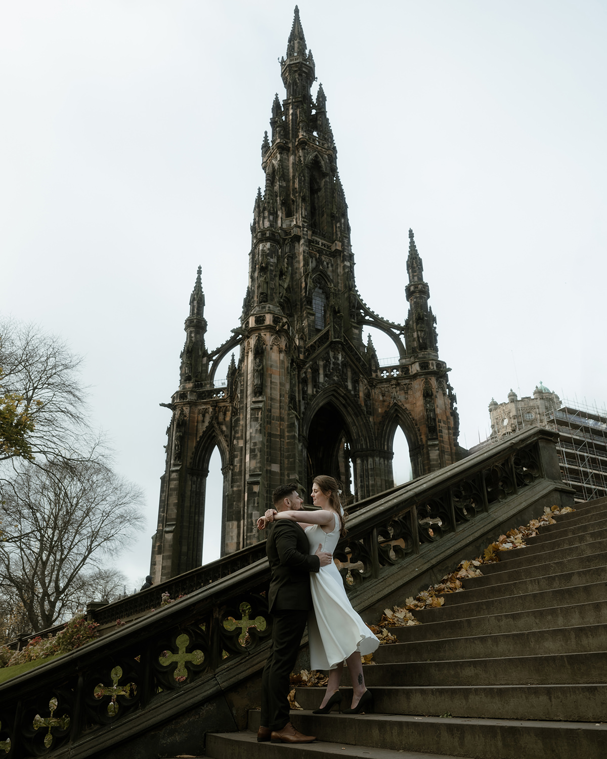 Home 17 Bride and groom embrace on a stone staircase beneath a towering Gothic Scott Monument in Edinburgh. The bride wears a sleeveless white dress and black heels, and the groom wears a dark suit. Fallen autumn leaves line the steps