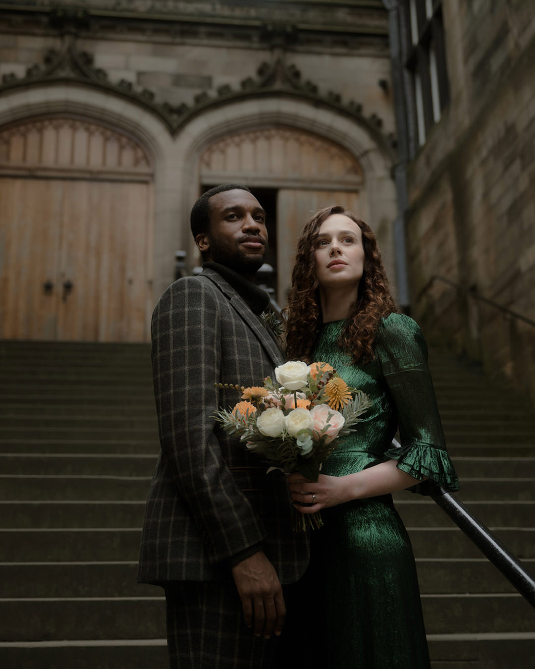 Home 18 Groom and bride stand on the steps of New College in Edinburgh. The bride wears a deep green dress and holds a bouquet of white and peach flowers, while the man wears a dark plaid coat. They gaze upward, framed by arched wooden doors and historic stone architecture on their elopement day in Edinburgh