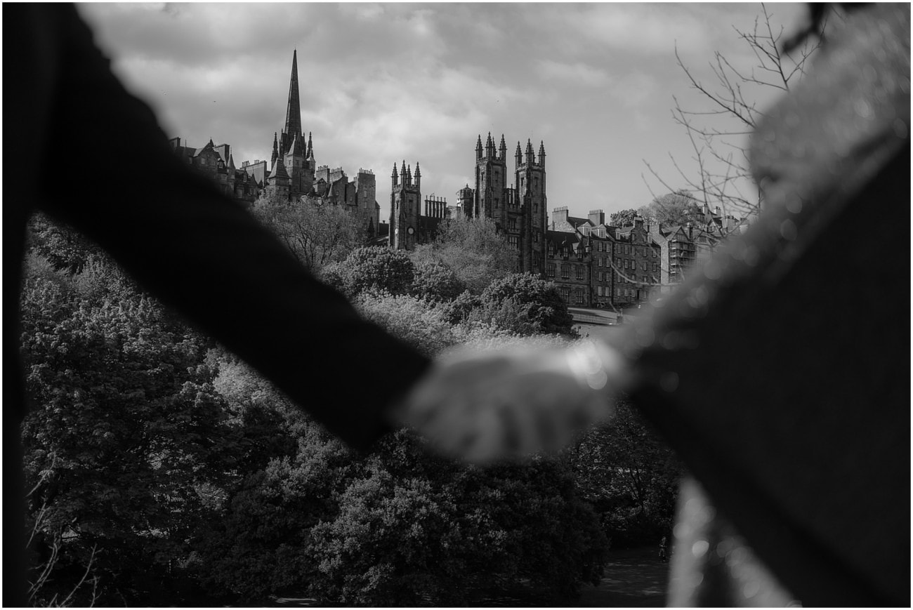 Black and white close-up of couple holding hands with gothic Edinburgh skyline and New College spire in the background