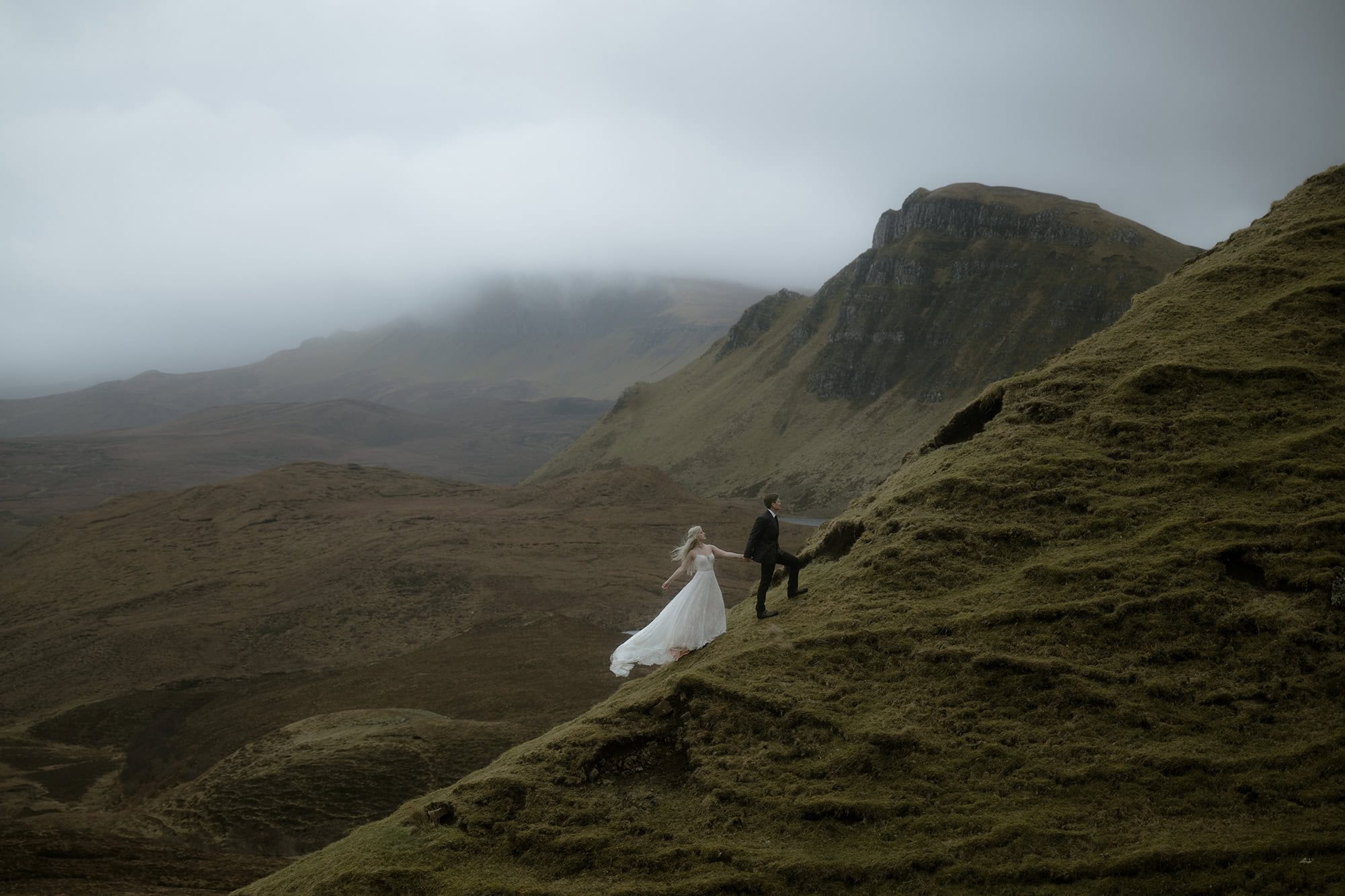 Couple climbing a mossy ridge on the Isle of Skye in wedding attire, dress billowing in the wind against a dramatic misty Highland landscape
