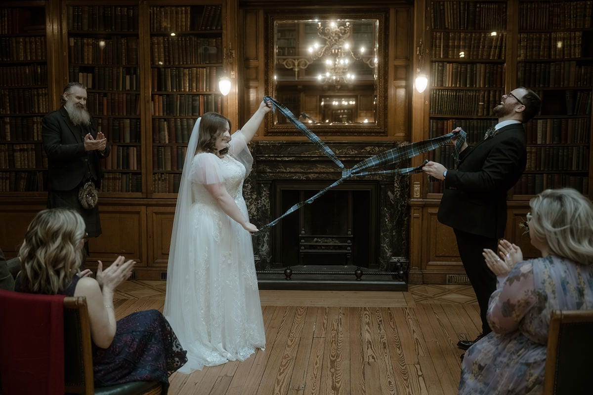 Bride and groom perform a tartan handfasting ceremony in the New Library at the Royal College of Physicians Edinburgh, surrounded by close family clapping, with antique bookshelves and fireplace behind them