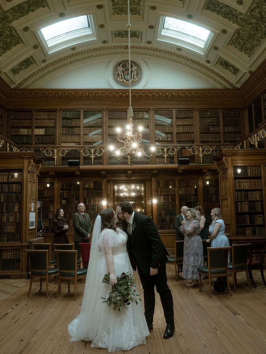 Bride and groom share their first kiss as a married couple in the New Library at the RCPE, with the full Georgian hall, ornate vaulted ceiling and chandelier visible behind them and guests celebrating in the background