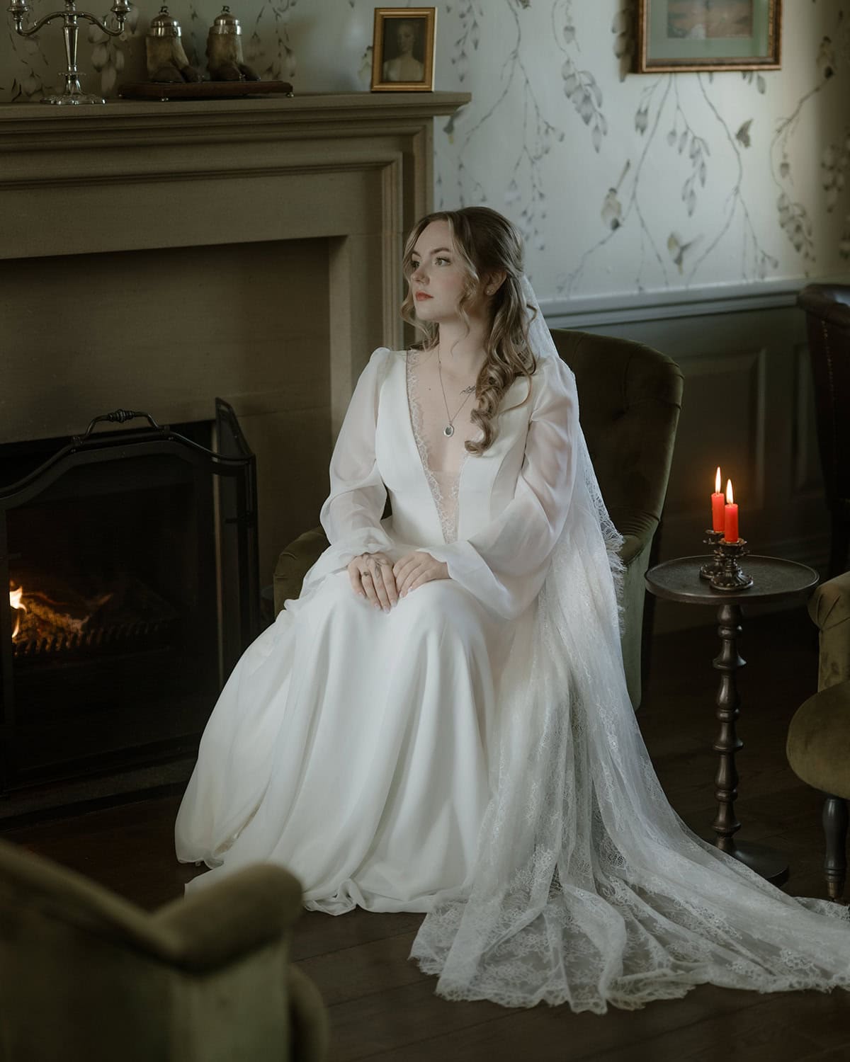 Bride in silk wedding dress seated by a lit fireplace with red candles at Monkstadt House on the Isle of Skye