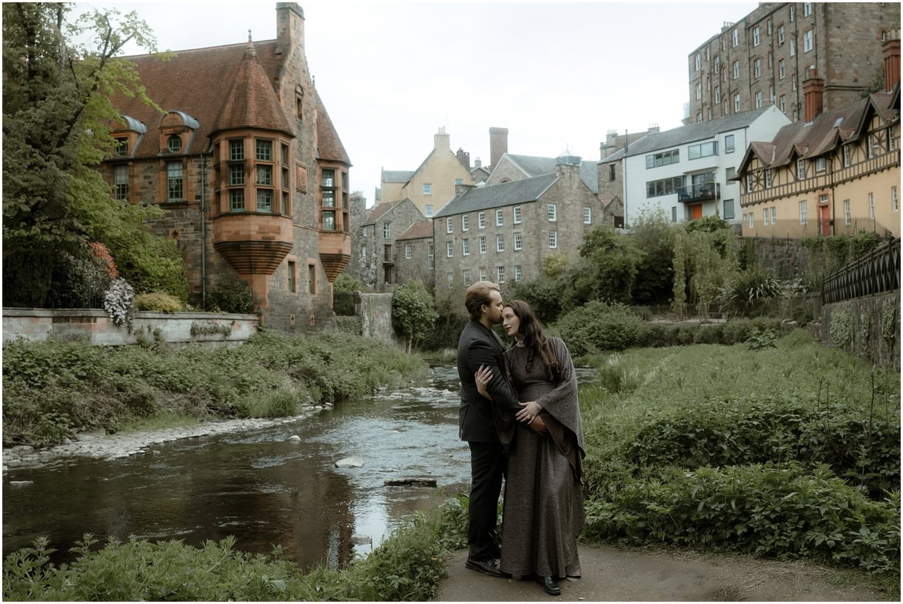 Couple embracing beside the Water of Leith with the full Well Court facade and Edinburgh skyline visible behind them