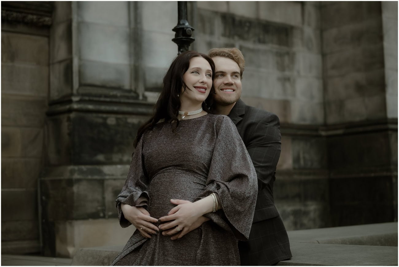 Couple smiling together with hands on baby bump outside St Giles Cathedral, dark academia maternity photography Edinburgh