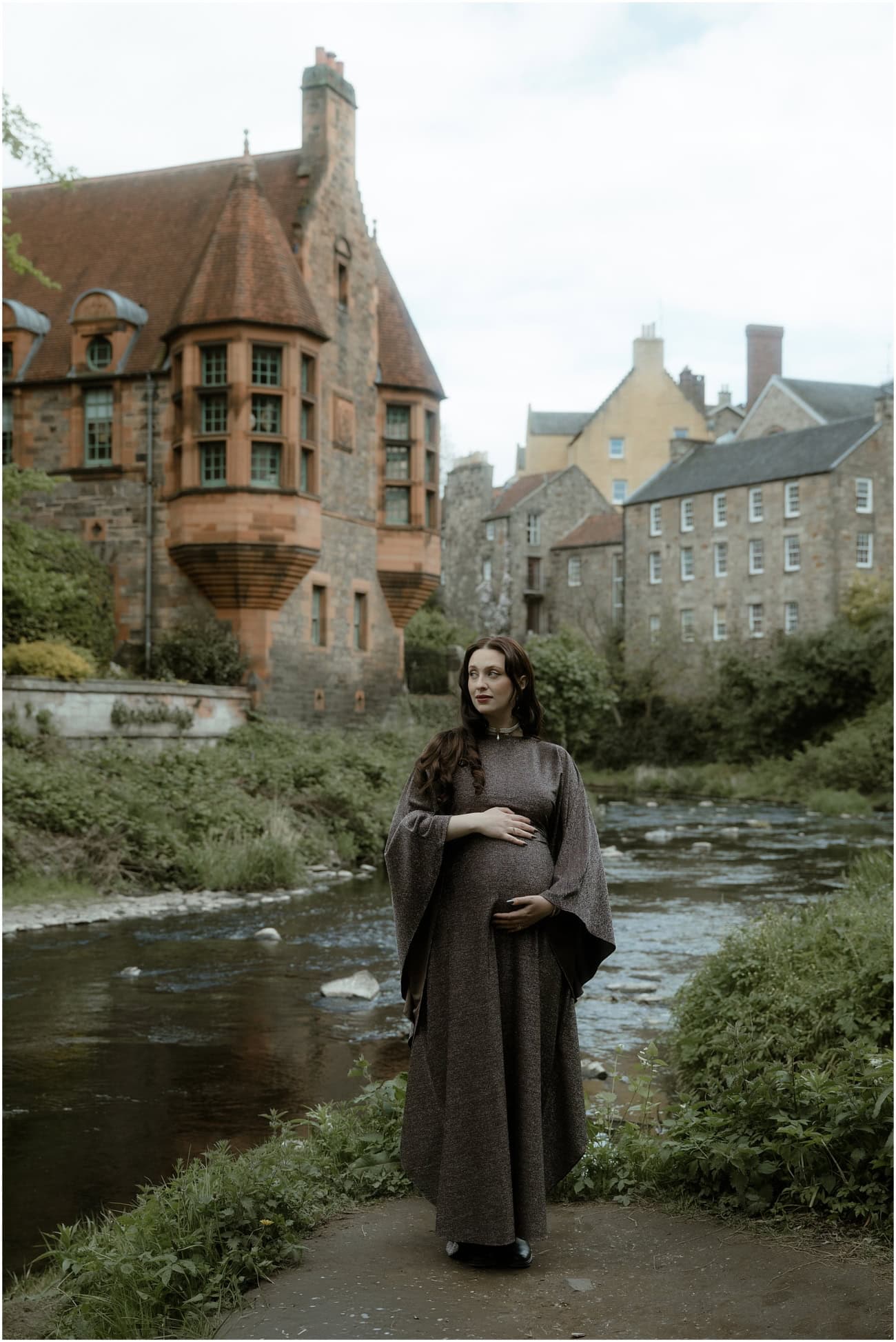 Solo maternity portrait of pregnant woman in a flowing dark cape holding her bump beside the Water of Leith with Well Court behind in Dean Village Edinburgh