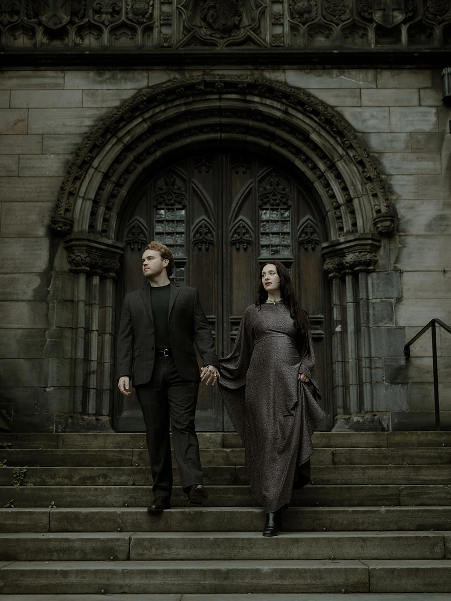 Couple walking down hand in hand on the steps of a grand gothic arched doorway of St Giles Cathedral in Edinburgh - atmospheric dark wedding portraits Scotland