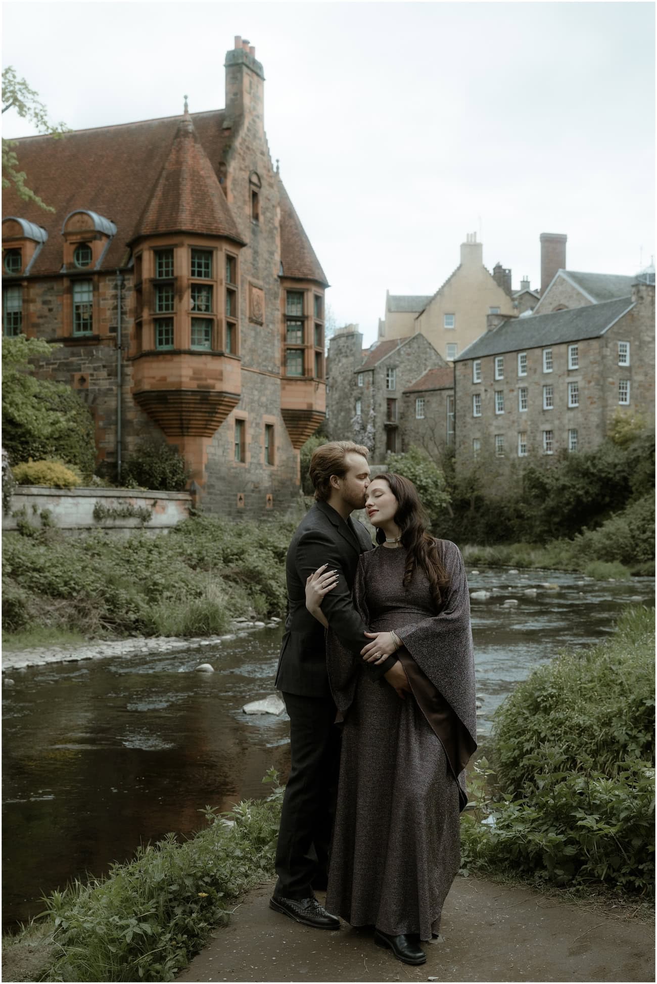 Couple embracing by the Water of Leith with the Well Court turret prominent behind during a dark academia Edinburgh maternity session