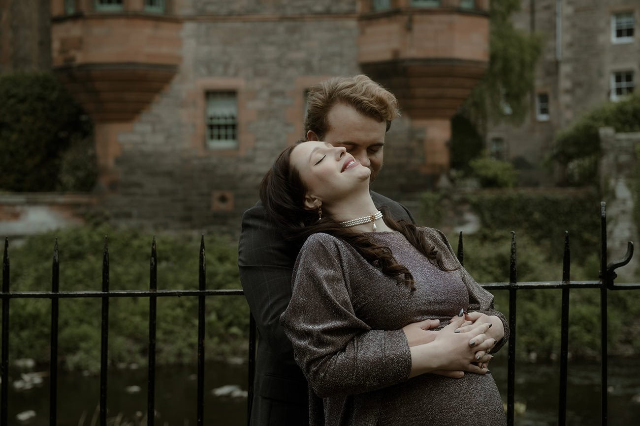 Couple sharing an intimate moment beside the Water of Leith at Dean Village Edinburgh, with the distinctive red stone Well Court building behind them
