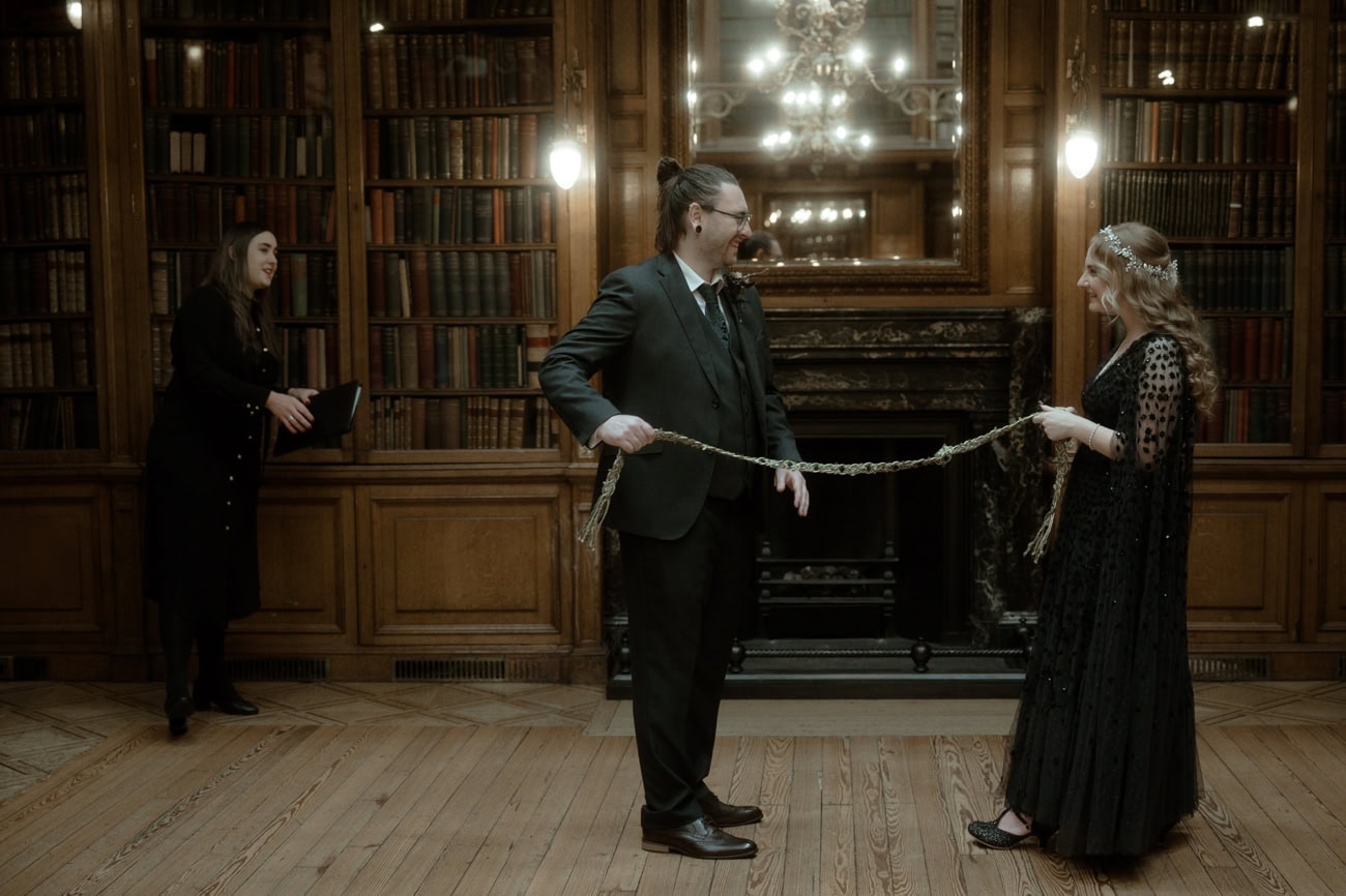 Weddings at the Royal College of Physicians Edinburgh: everything you need to know 12 Couple performing handfasting ceremony in the New Library at the Royal College of Physicians Edinburgh with celebrant and Georgian fireplace