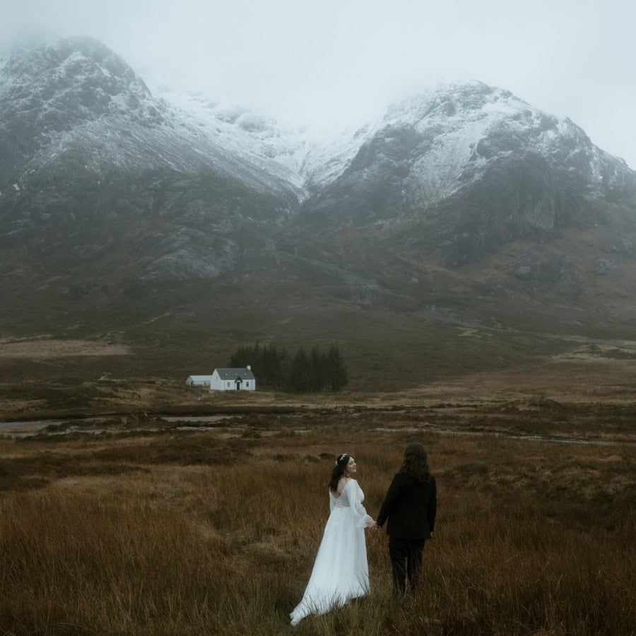 Bride and groom walking hand in hand through Glencoe moorland with snow-dusted mountains and low cloud - moody elopement photos Scotland