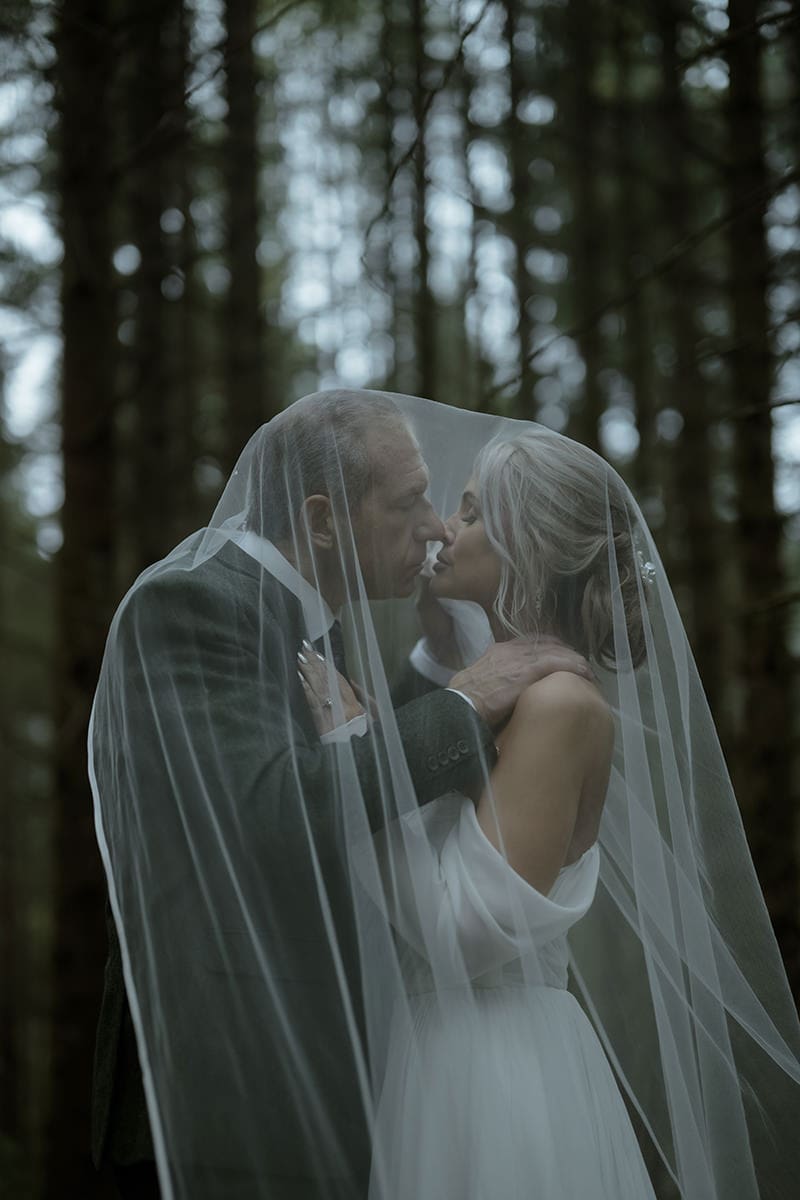 Bride and groom kissing beneath a flowing veil in a dark pine forest of Glencoe Lochan