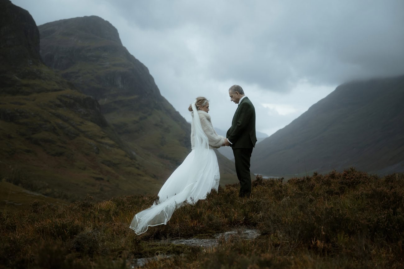 Bride and groom holding hands on the Glencoe moorland in October, wedding dress and veil billowing in the wind against dramatic cloud-covered mountains