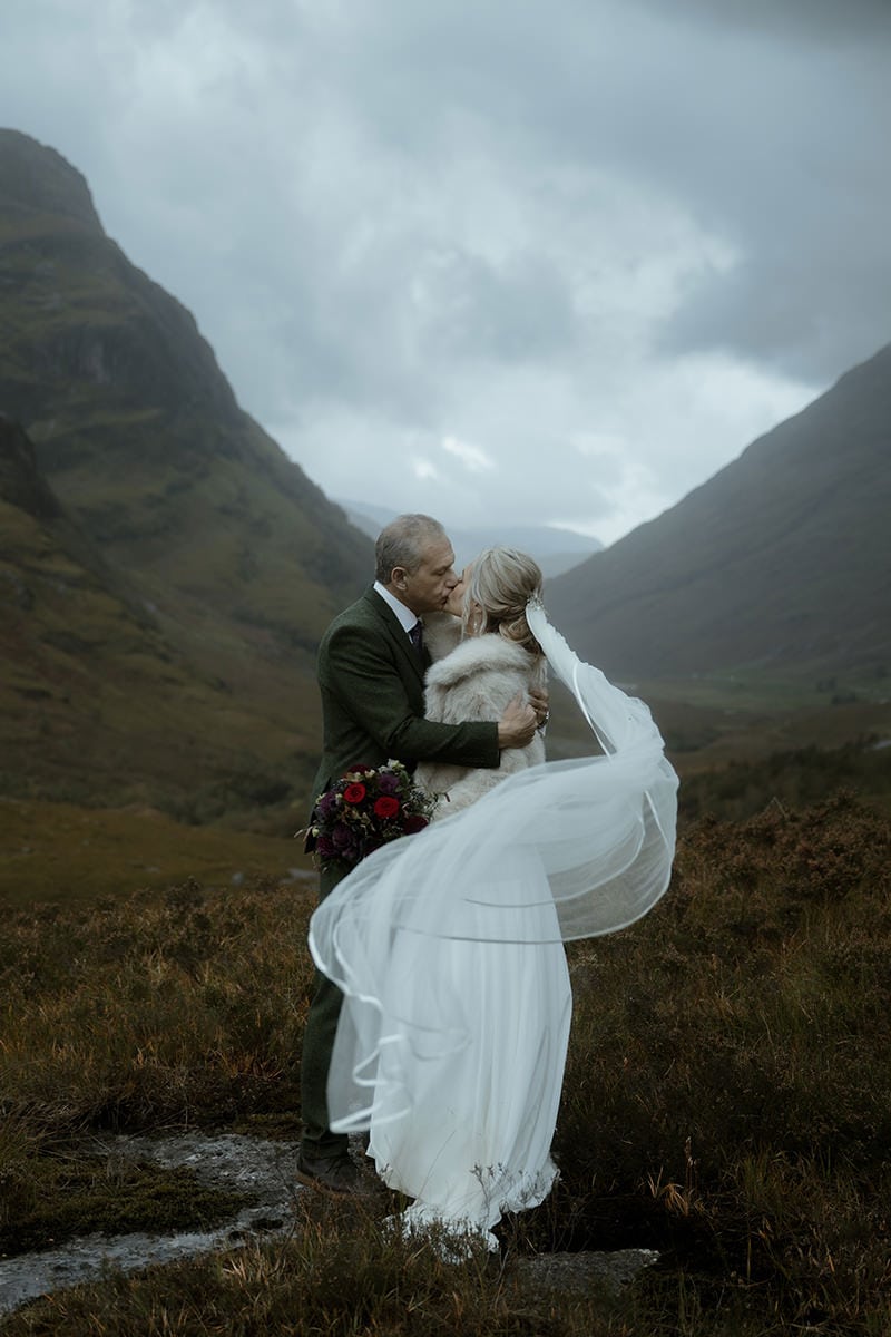 Bride and groom kissing in Glencoe with the wedding veil sweeping dramatically in the wind, moody dark mountains and overcast sky behind them — cinematic Scotland elopement