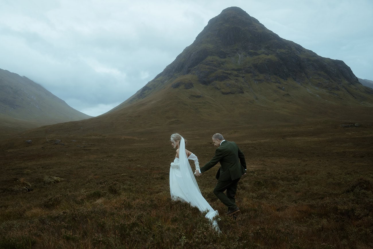 Bride and groom walking hand in hand across the Glencoe moorland toward a dramatic highland peak on an overcast autumn day
