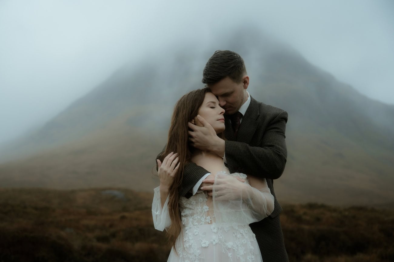 Groom tenderly holding bride's face as they embrace in Glencoe, a fog-covered mountain rising dramatically behind them - moody elopement photography Scotland