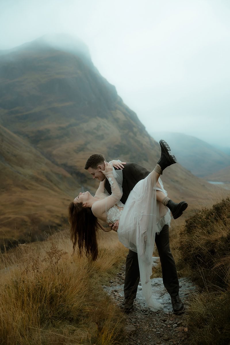 Groom lifting bride on a misty Glencoe hillside trail, both laughing, with dramatic fog-covered mountains in the background - cinematic Scotland elopement photos