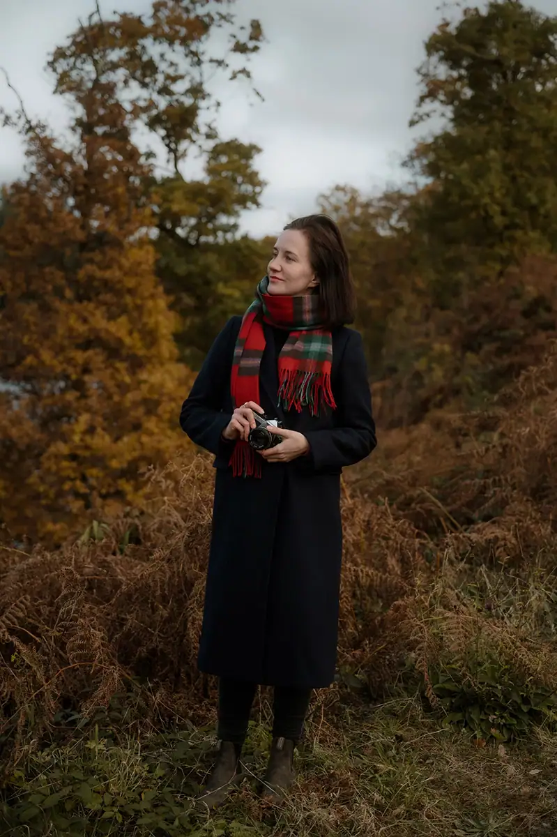 Edinburgh Elopement Photographer 1 Natalia Swiader, Edinburgh elopement photographer, standing in autumn woodland holding a film camera, wearing a tartan scarf