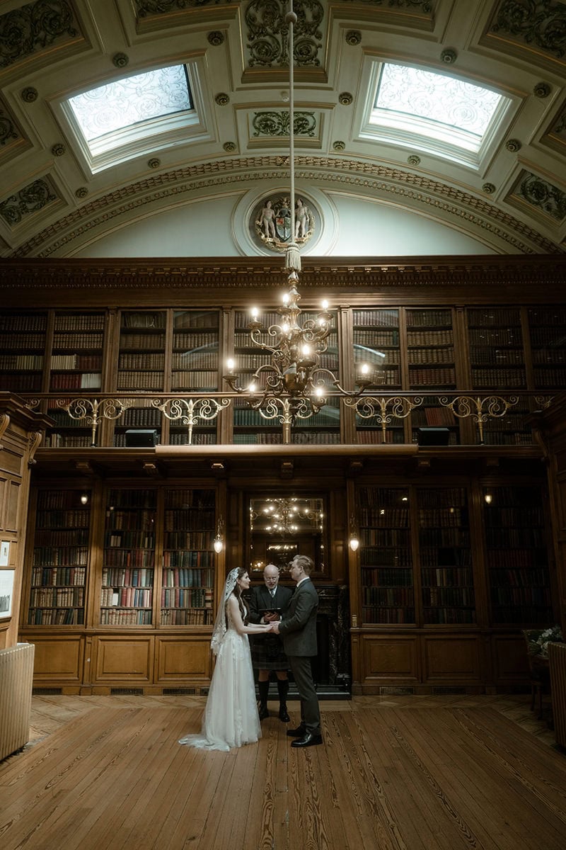 Bride and groom exchanging vows in the New Library at the Royal College of Physicians of Edinburgh, surrounded by floor-to-ceiling bookshelves and a grand chandelier - intimate Edinburgh elopement venue