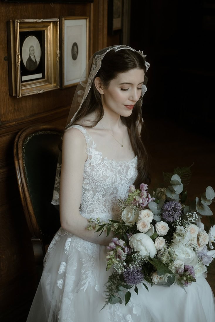 Bride in lace wedding dress and floral veil seated in a dark wood-panelled room at the Royal College of Physicians of Edinburgh, holding a bouquet of roses and alliums