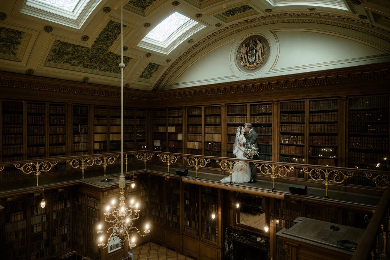 Bride and groom sharing their first kiss on the gallery balcony of the New Library at RCPE Edinburgh, the grand two-storey library and ornate chandelier below them