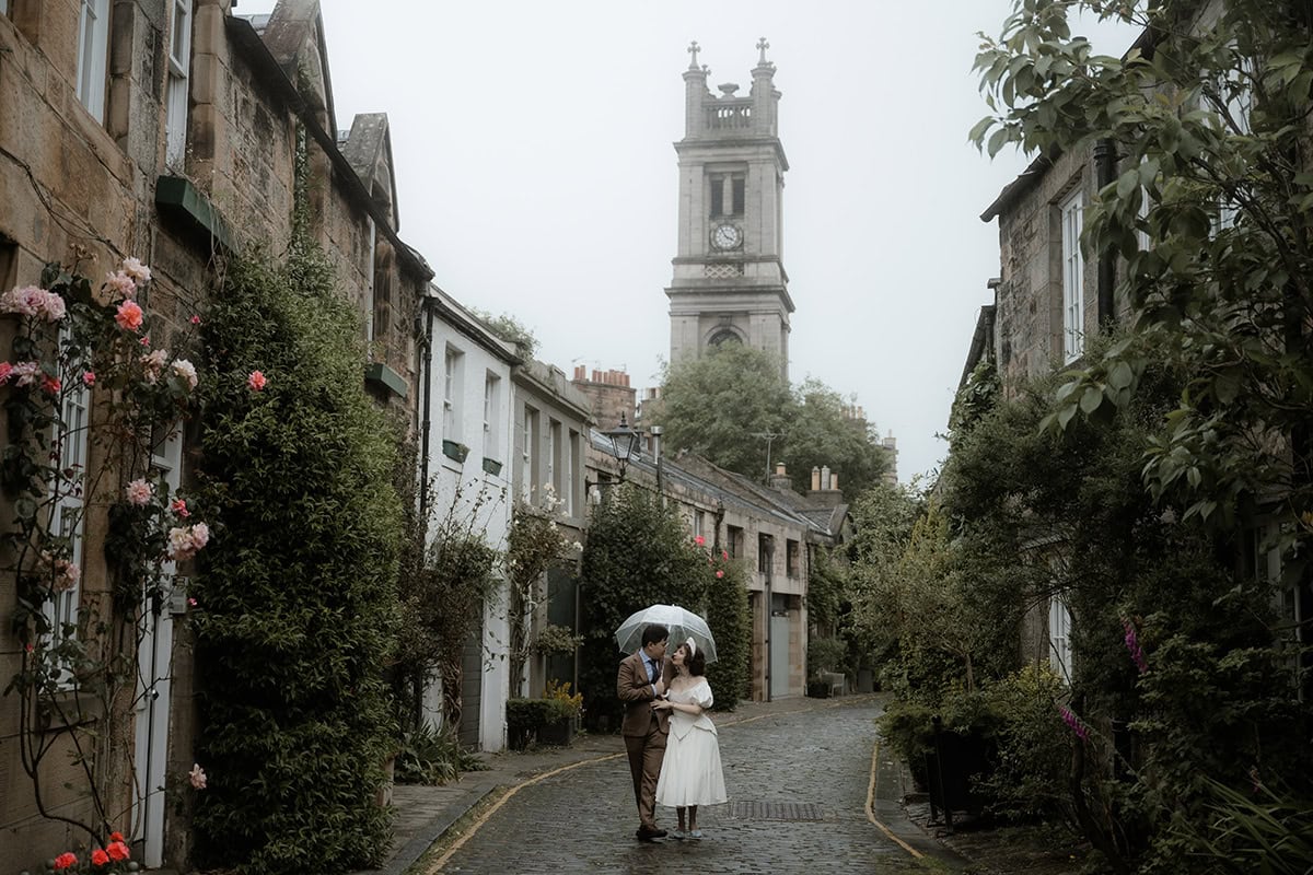 Bride and groom share a kiss under a clear umbrella on a rainy day in Circus Lane, Stockbridge, Edinburgh, surrounded by rose-covered stone cottages and St Stephen's Church tower rising through the mist