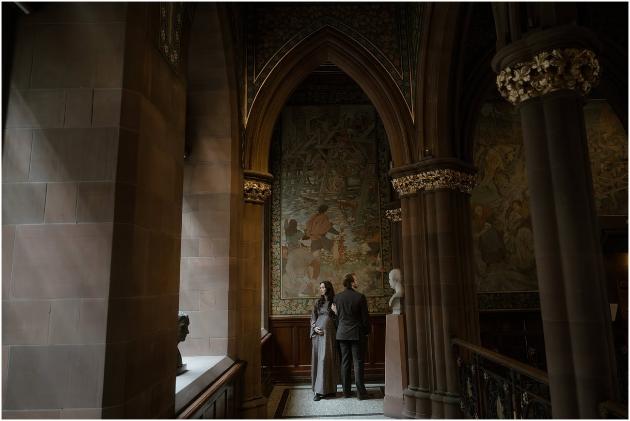 Couple standing small beneath a soaring gothic arch and historic mural in the grand interior of the Scottish National Portrait Gallery Edinburgh
