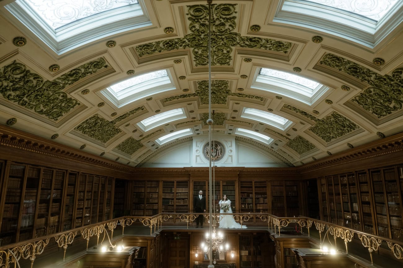 Bride and groom standing on the gallery level of the New Library at the Royal College of Physicians Edinburgh, looking down into the grand Georgian hall with ornate vaulted ceiling and floor-to-ceiling bookshelves