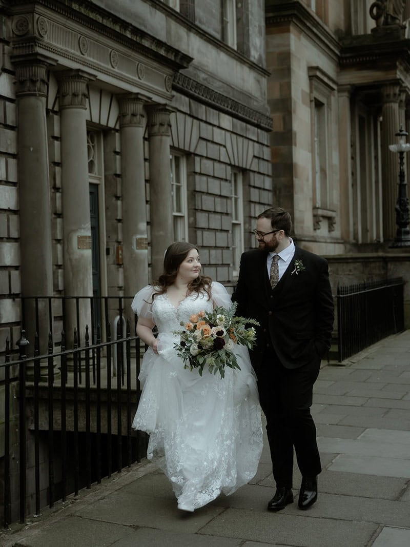 Bride in a tulle puff-sleeve gown and groom in a dark suit walk together laughing along a Georgian street in Edinburgh New Town after their Royal College of Physicians wedding