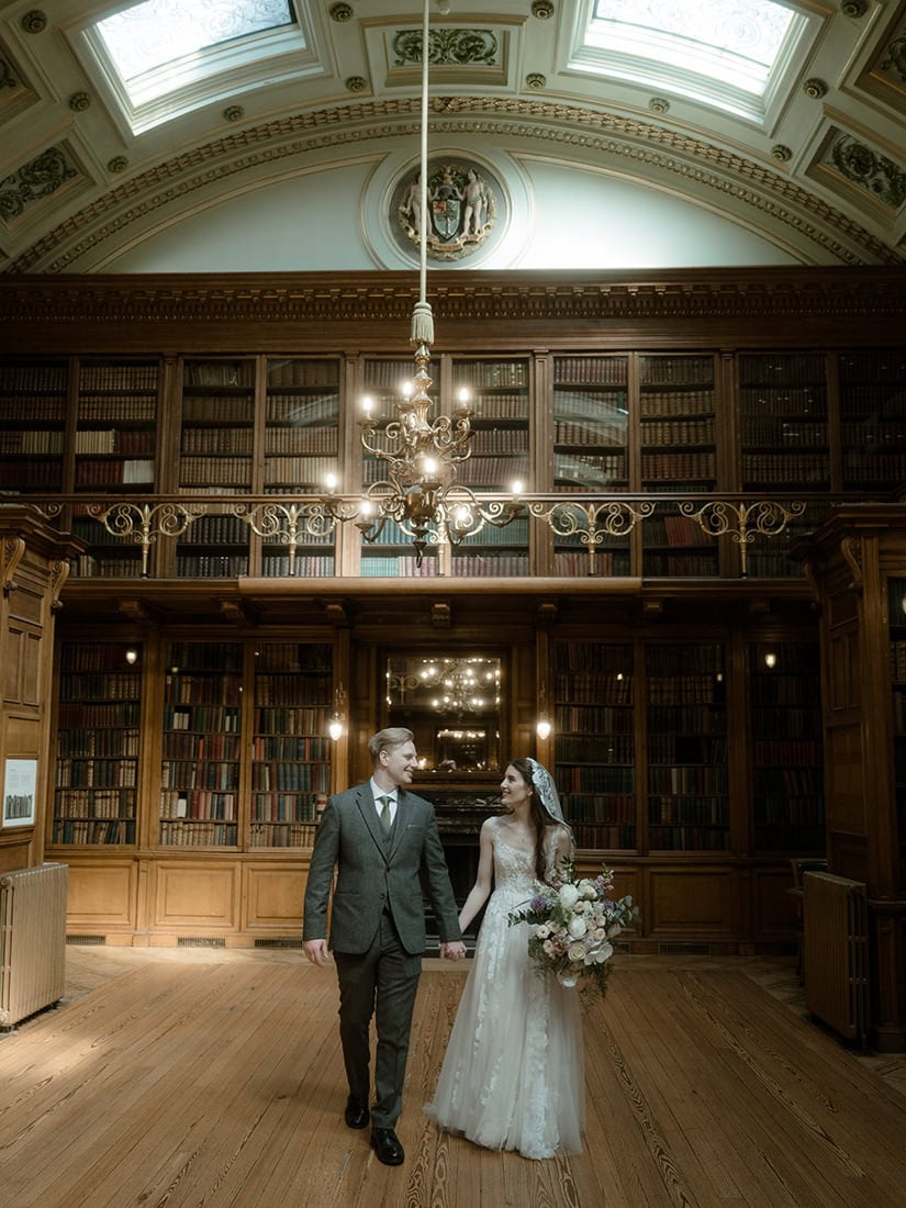 Weddings at the Royal College of Physicians Edinburgh: everything you need to know 20 Bride and groom walking hand in hand in the New Library at the Royal College of Physicians Edinburgh, floor-to-ceiling bookshelves and chandelier