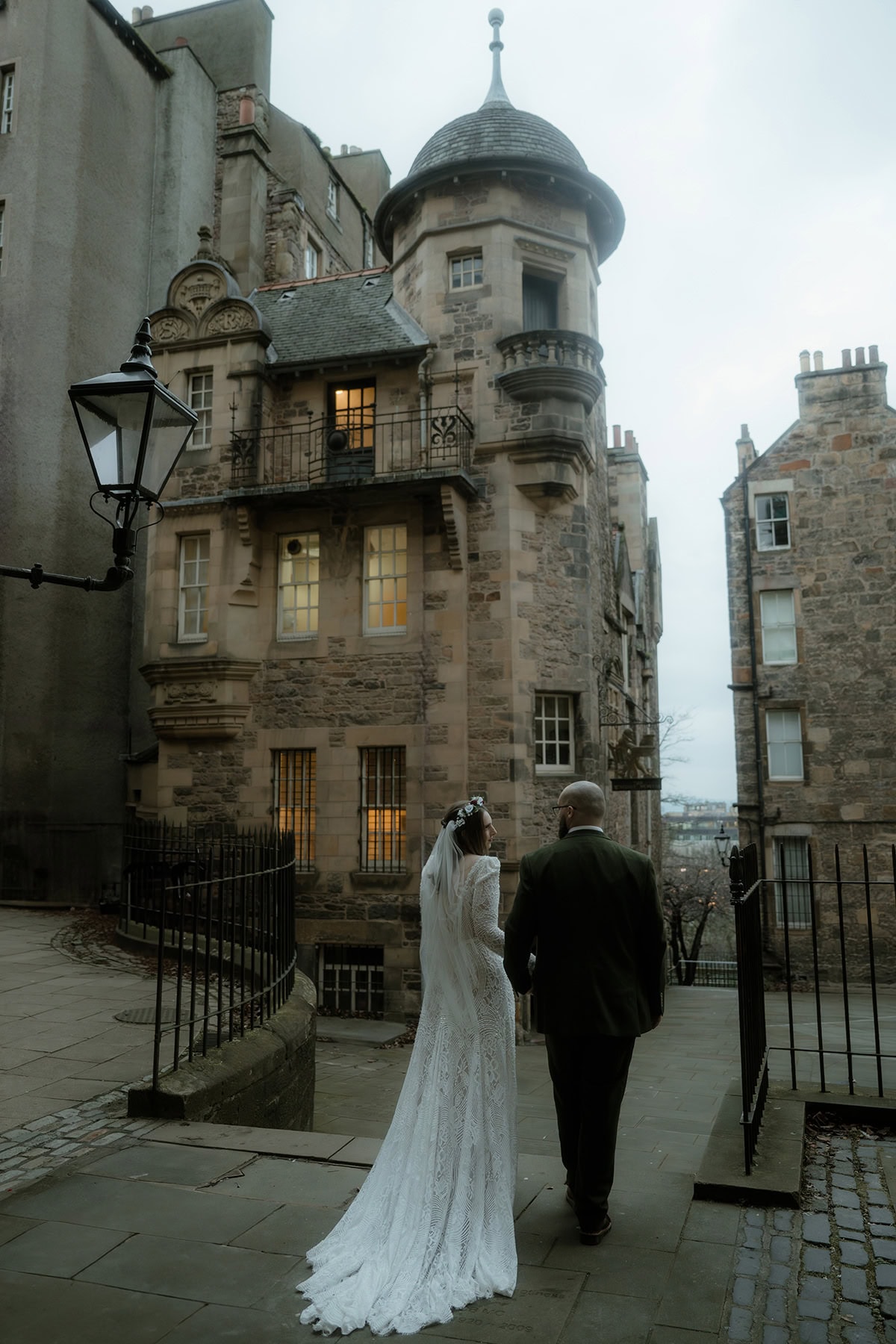 Bride and groom walking hand in hand through Edinburgh's Old Town at dusk in winter, the Writers' Museum turret glowing warmly behind them
