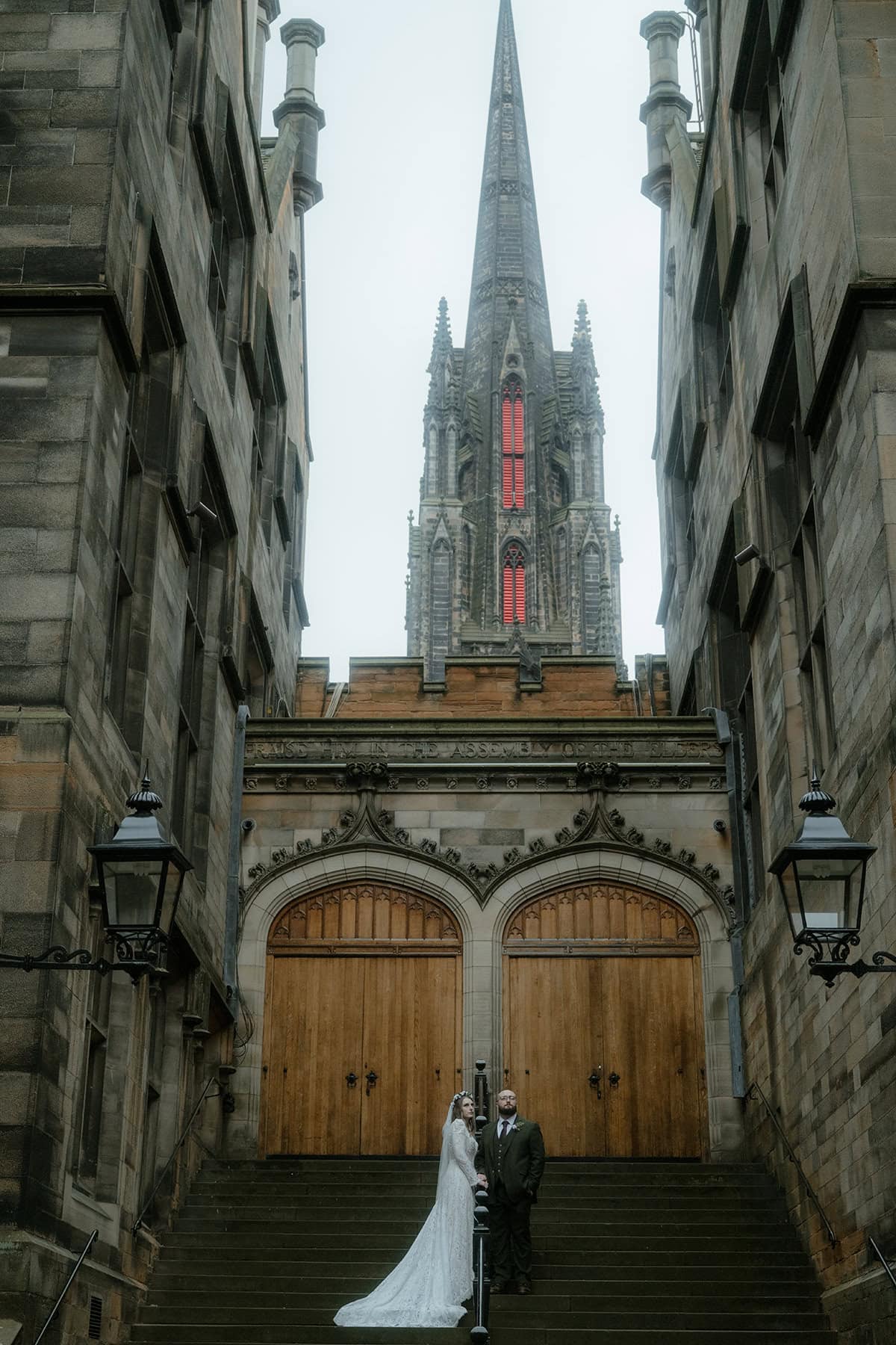 Bride and groom standing at the top of the New College steps in Edinburgh, dwarfed by the dramatic gothic spire above them